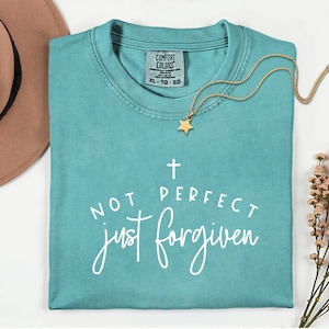 May include: A turquoise Comfort Colors t-shirt with the text "NOT PERFECT just forgiven" in white script. A gold necklace with a star pendant rests on the shirt. A brown hat is visible in the upper left corner.