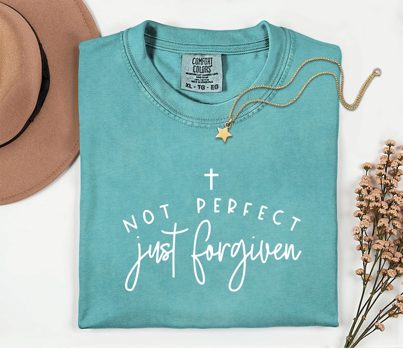May include: A turquoise Comfort Colors t-shirt with the text "NOT PERFECT just forgiven" in white script. A gold necklace with a star pendant rests on the shirt. A brown hat is visible in the upper left corner.