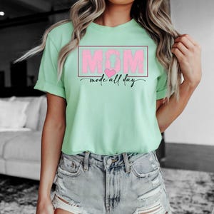 Puede incluir: Camiseta verde claro con la palabra "MOM" en letras rosas dentro de un rect&aacute;ngulo rosa, con un coraz&oacute;n rosa y las palabras "mode all day" en cursiva. Se combina con pantalones cortos de mezclilla.