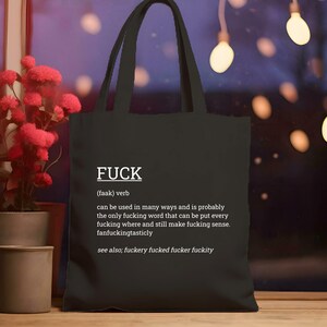 May include: Black tote bag with the word "FUCK" in white, along with a definition and related terms. The bag has two handles and is displayed in front of a window with string lights and a plant.