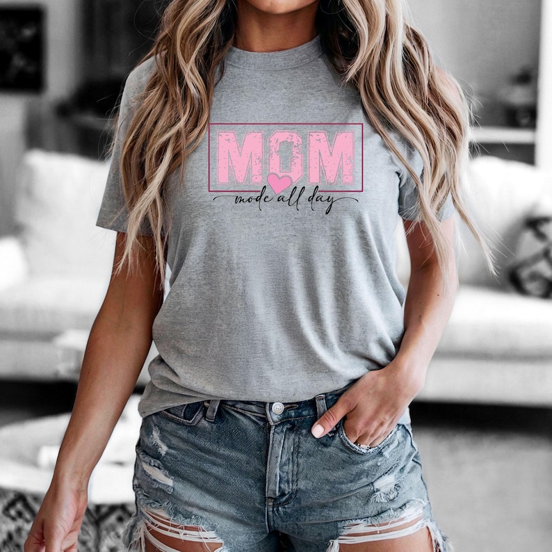Puede incluir: Una camiseta gris con la palabra "MOM" en un recuadro rosa, un coraz&oacute;n rosa y la frase "mode all day" en letra cursiva. La camiseta es de manga corta.