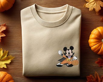 May include: A beige crewneck sweatshirt featuring a small graphic of Mickey Mouse dressed in a vampire costume with a cape and orange suit, holding a jack-o'-lantern bucket. The character is positioned on the lower right side of the garment.