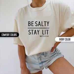 May include: A white t-shirt with the text "BE SALTY STAY LIT" and the scripture reference "MATT 5:13-14" printed in black. The shirt is being worn by a person with a light tan complexion. The person is wearing denim shorts.