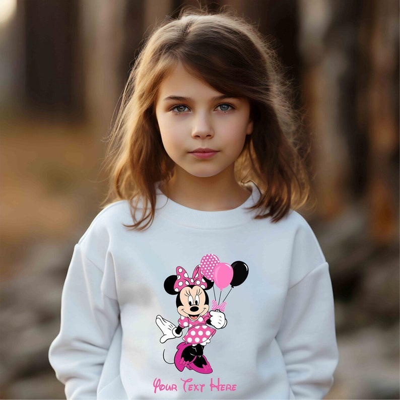 Puede incluir: Sudadera blanca con un gr&aacute;fico rosa de Minnie Mouse sosteniendo globos rosas y negros. El texto "Your Text Here" est&aacute; impreso debajo del gr&aacute;fico.