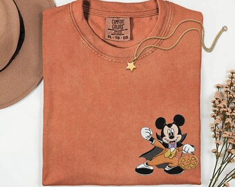 May include: A rust-colored short-sleeve t-shirt featuring a graphic of Mickey Mouse dressed as a vampire holding a jack-o'-lantern. The shirt has a crew neckline and a Comfort Colors brand label printed on the inside collar with size XL listed.