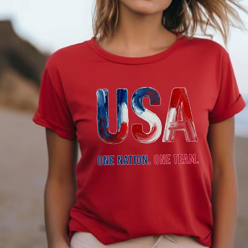 K&ouml;nnte beinhalten: Rotes T-Shirt mit dem Wort "USA" in Blau und Rot sowie dem Text "ONE NATION. ONE TEAM." Das Shirt hat kurze &Auml;rmel und einen Rundhalsausschnitt.