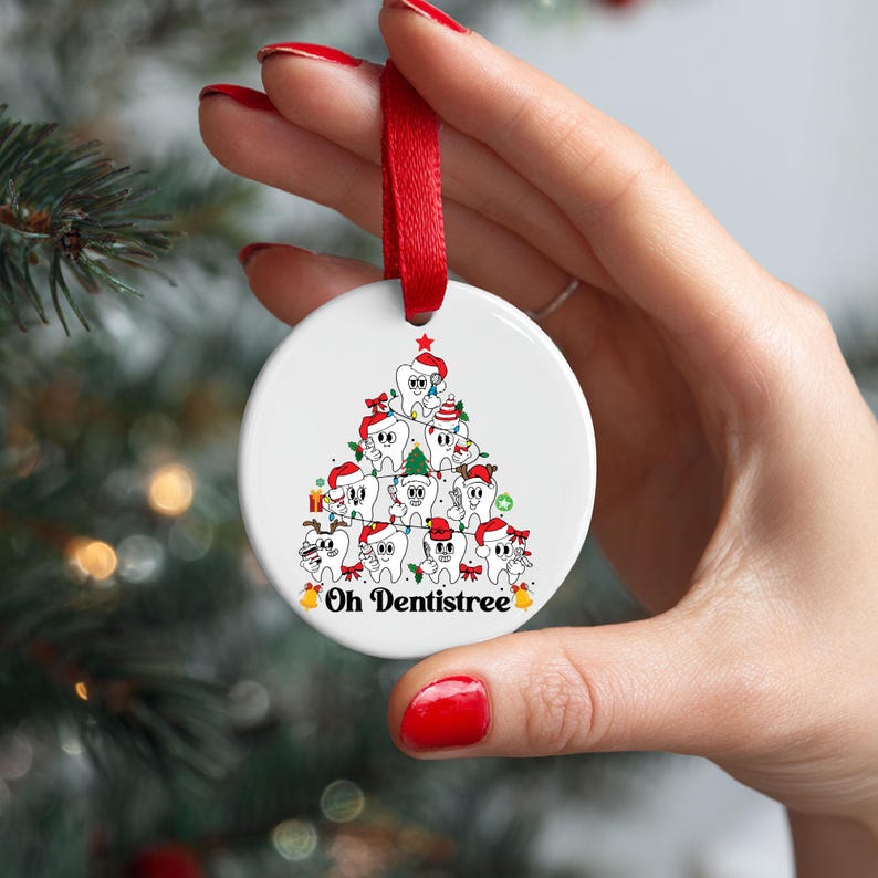 Puede incluir: Un adorno navide&ntilde;o de cer&aacute;mica blanca con una cinta roja. El adorno presenta un &aacute;rbol de Navidad de dibujos animados hecho de dientes sonrientes con gorros de Pap&aacute; Noel. El texto "Oh Dentistree" est&aacute; en la parte inferior.