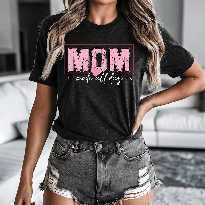 Puede incluir: Camiseta gris oscuro con la palabra "MOM" en letras rosas dentro de un rect&aacute;ngulo rosa, con un coraz&oacute;n rosa debajo. La frase "mode all day" est&aacute; escrita en cursiva debajo del coraz&oacute;n. La camiseta se usa con pantalones cortos de mezclilla grises.