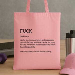 May include: A pink tote bag with the word "FUCK" printed in black, along with a definition. The bag is made of a soft material and has handles for carrying. A pink baseball cap and a water bottle are in the background.