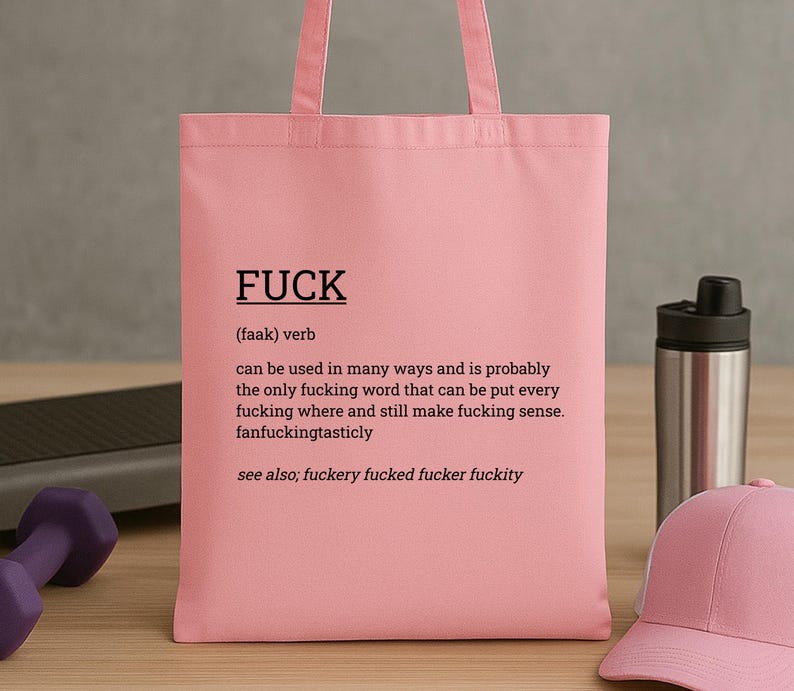May include: A pink tote bag with the word "FUCK" printed in black, along with a definition. The bag is made of a soft material and has handles for carrying. A pink baseball cap and a water bottle are in the background.