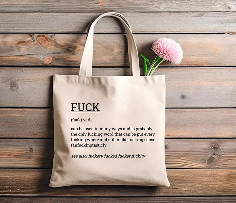 May include: Cream-colored tote bag with the word "FUCK" printed in black, along with a definition. The bag has a simple design with long handles. A pink flower with green stems is placed inside the bag.