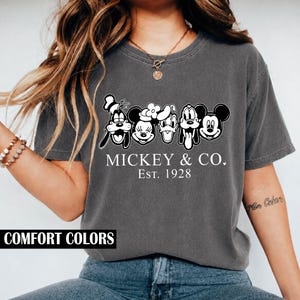 Pode incluir: Uma t-shirt de manga curta cinzento-carvão com uma impressão gráfica branca de cinco personagens de desenhos animados alinhados. Abaixo das personagens, o texto diz MICKEY & CO. EST. 1928. A t-shirt tem gola redonda e um corte descontraído.