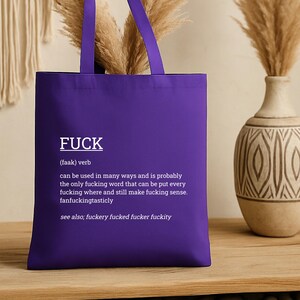 May include: A purple tote bag with the word "FUCK" in white, along with a definition. The bag has long straps and is made of a sturdy material. The background includes a vase and decorative elements.