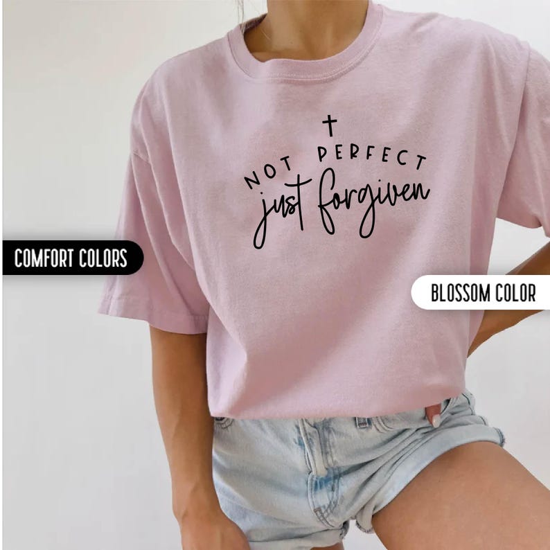 May include: A light pink t-shirt with the text "NOT PERFECT just forgiven" in black script. The shirt has short sleeves and a crew neck. The shirt is a blossom color.