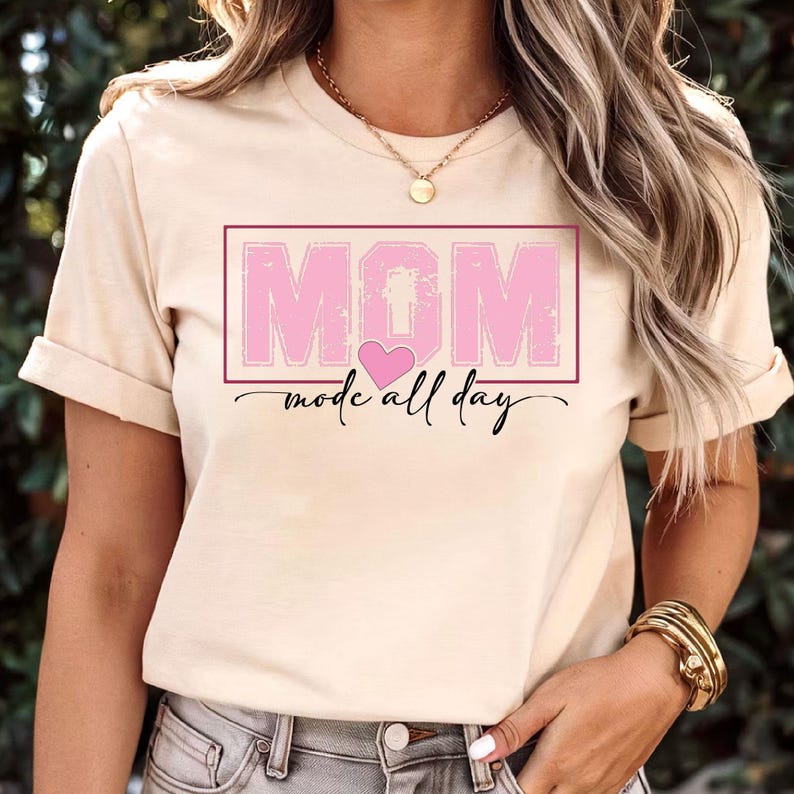 Puede incluir: Camiseta beige con la palabra "MOM" en letras rosas dentro de un rect&aacute;ngulo rosa, con un coraz&oacute;n rosa y la frase "mode all day" en cursiva. La camiseta tiene las mangas dobladas.