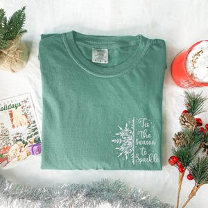 Pode incluir: Uma t-shirt de manga curta verde-sálvia com um gráfico branco no lado inferior direito, apresentando um design de floco de neve e o texto 'Tis the Season to Sparkle'. A t-shirt tem gola redonda e uma etiqueta Comfort Colors no interior.