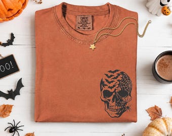 May include: A burnt orange short-sleeve t-shirt featuring a black graphic of a skull on the lower right side. The skull design incorporates a spider web, a spider, and a flock of bats. The shirt has a crew neckline and a Comfort Colors label.