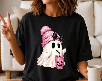 May include: A black t-shirt featuring a graphic of a white ghost wearing a pink knit beanie with a pom-pom and a black bow. The ghost is holding a pink cup with a jack-o'-lantern face and a black and white striped straw, drinking from it.
