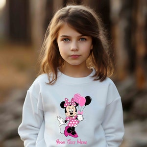 Puede incluir: Sudadera blanca con un gr&aacute;fico de Minnie Mouse rosa y negro sosteniendo globos rosas y negros. El texto "Your Text Here" est&aacute; impreso debajo del gr&aacute;fico.