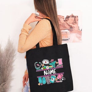 May include: Black tote bag with a colorful design of medical symbols and the word "NAME" in the center. The design includes a stethoscope, pills, a syringe, a bandage, and a heart.