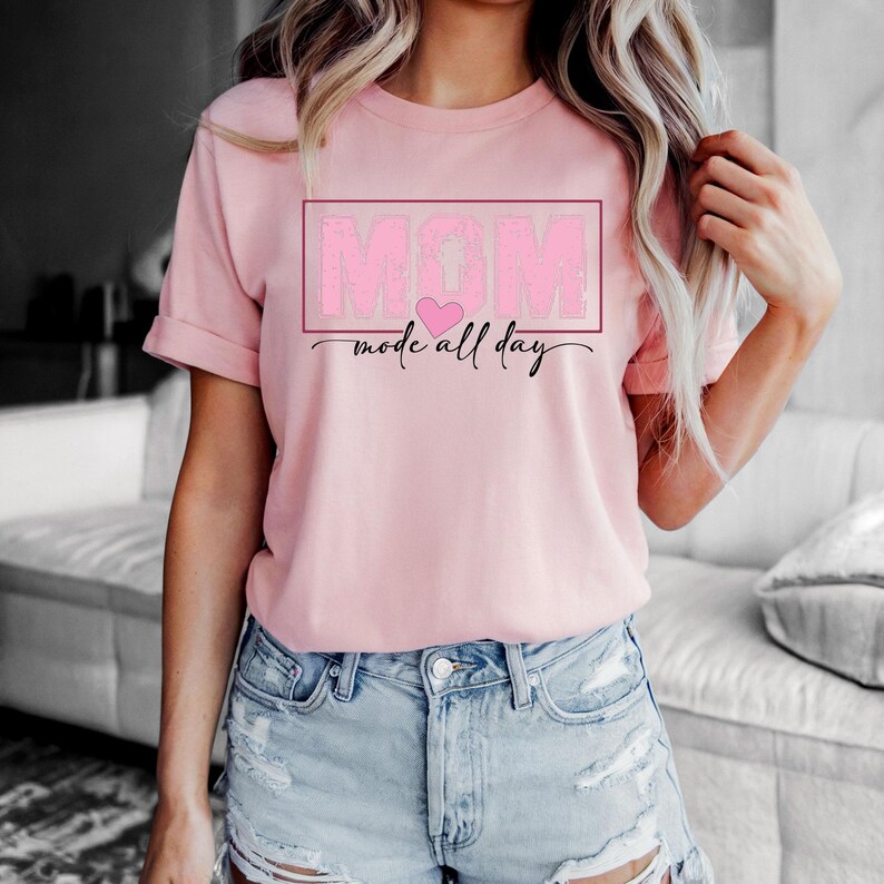 Puede incluir: Una camiseta rosa claro con la palabra "MOM" en un marco rectangular rosa, un coraz&oacute;n rosa y la frase "mode all day" en cursiva. La camiseta es de cuello redondo y se est&aacute; usando.