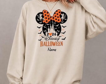 May include: A beige crewneck sweatshirt featuring a graphic of Minnie Mouse ears with an orange polka dot bow, surrounded by black bats. The text reads 2026 Disney HALLOWEEN Name, with a small orange Mickey Mouse icon replacing the zero in 2026.