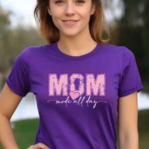 Puede incluir: Una camiseta morada con la palabra "MOM" en letras rosas desgastadas dentro de un rect&aacute;ngulo rojo. Debajo de la palabra "MOM" est&aacute; la frase "mode all day" con un coraz&oacute;n rosa.