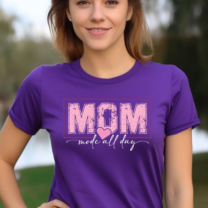 Puede incluir: Una camiseta morada con la palabra "MOM" en letras rosas desgastadas dentro de un rect&aacute;ngulo rojo. Debajo de la palabra "MOM" est&aacute; la frase "mode all day" con un coraz&oacute;n rosa.