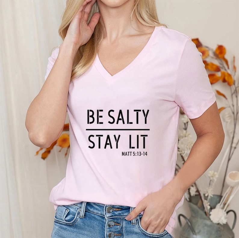 May include: A pink v-neck t-shirt with the text "BE SALTY STAY LIT" and "MATT 5:13-14" printed on the front.