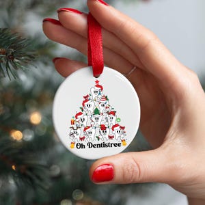 Puede incluir: Adorno navide&ntilde;o redondo y blanco con una cinta roja. Presenta un &aacute;rbol de Navidad de dibujos animados hecho de dientes con gorros de Pap&aacute; Noel. El texto "Oh Dentistree" est&aacute; impreso en la parte inferior.