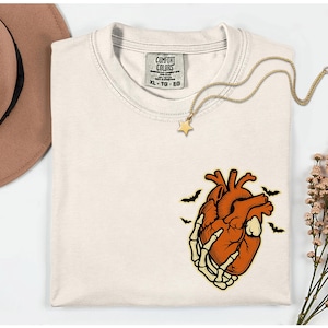 May include: Cream-colored t-shirt with a graphic of an anatomical heart held by a skeleton hand, with bats. A gold star pendant necklace is draped across the shirt. The shirt has a Comfort Colors tag.