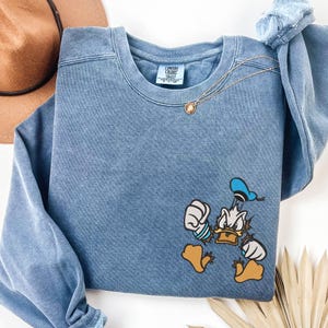 Pode incluir: Uma camisola azul de gola redonda com um gráfico do Pato Donald no lado inferior direito. A camisola tem um aspeto desbotado de estilo vintage e uma etiqueta da marca Comfort Colors no interior da gola. Uma corrente dourada fina repousa sobre o decote.