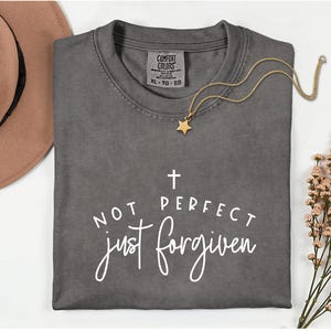 May include: A gray Comfort Colors t-shirt with the text "NOT PERFECT just forgiven" in white script. A gold star pendant necklace rests on the shirt. A brown hat and flowers are in the background.