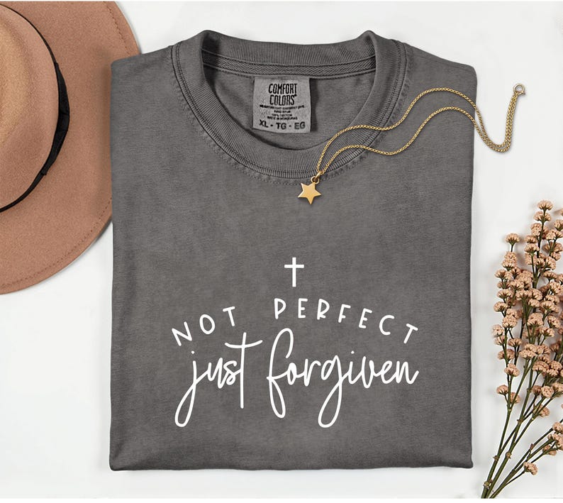 May include: A gray Comfort Colors t-shirt with the text "NOT PERFECT just forgiven" in white script. A gold star pendant necklace rests on the shirt. A brown hat and flowers are in the background.