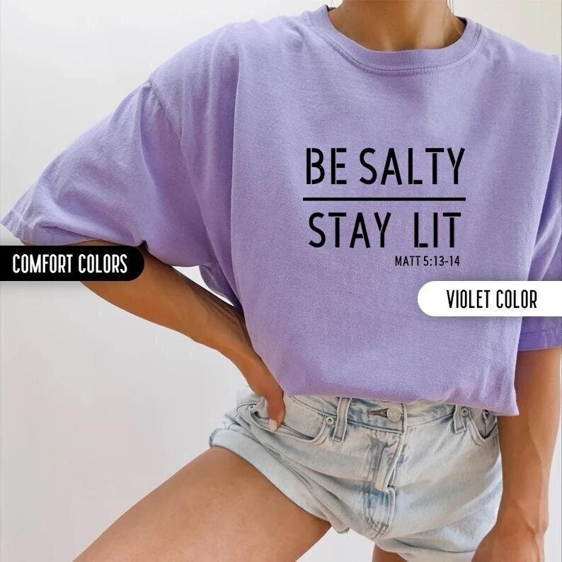 May include: A purple t-shirt with the text "BE SALTY STAY LIT" and the scripture reference "MATT 5:13-14" printed on the front. The shirt is made of a soft, comfortable material.