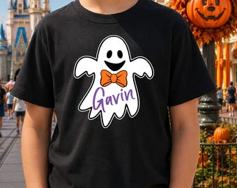 May include: A black short-sleeved t-shirt featuring a white ghost graphic centered on the chest. The ghost has a smiling face, an orange bow tie, and the name Gavin written in purple script lettering below the bow tie on the ghost's body.