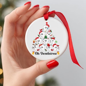 Puede incluir: Adorno navide&ntilde;o redondo de cer&aacute;mica blanca con cinta roja. Presenta un dise&ntilde;o de &aacute;rbol de Navidad hecho de dientes de dibujos animados con gorros y bufandas de Pap&aacute; Noel. El texto "Oh Dentistree" est&aacute; en la parte inferior.