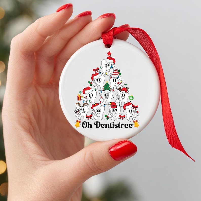 Puede incluir: Adorno navide&ntilde;o redondo de cer&aacute;mica blanca con cinta roja. Presenta un dise&ntilde;o de &aacute;rbol de Navidad hecho de dientes de dibujos animados con gorros y bufandas de Pap&aacute; Noel. El texto "Oh Dentistree" est&aacute; en la parte inferior.