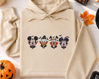 May include: A beige hooded sweatshirt featuring an embroidered graphic of four cartoon characters dressed as vampires. The design includes Mickey Mouse, Donald Duck, Pluto, and Minnie Mouse, each wearing a cape, surrounded by small black bats.