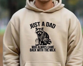Könnte beinhalten: Ein beiger Hoodie mit einem schwarzen Aufdruck eines Waschbären, der eine Milchpackung hält. Der Text lautet "Just a Dad who always came back with the milk."
