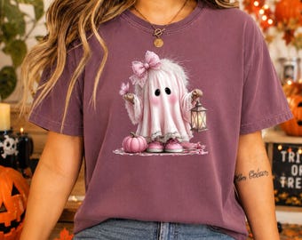 May include: A mauve short-sleeve t-shirt featuring a graphic of a ghost wearing a pink bow and sneakers, holding a lantern next to a small pink pumpkin. The ghost has large black eyes and rosy cheeks, with a butterfly resting on its hand.