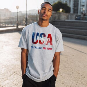 K&ouml;nnte beinhalten: Hellgraues T-Shirt mit dem Wort "USA" in rot, wei&szlig; und blau. Darunter steht der Text "ONE NATION. ONE TEAM.". Das T-Shirt hat kurze &Auml;rmel.