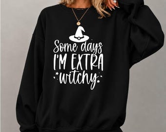 May include: A black long-sleeve crewneck sweatshirt featuring a white graphic print of a witch hat and the text Some days I'M EXTRA witchy with small star accents. The design is centered on the chest of the garment, which has a relaxed fit.