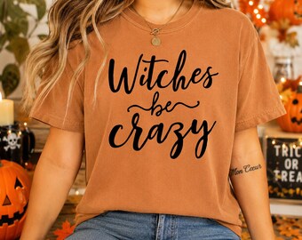 May include: A burnt orange short-sleeve t-shirt featuring the black text Witches be crazy printed in a cursive script font across the chest. The garment has a crew neckline and a relaxed fit, showcasing a solid color throughout the fabric.