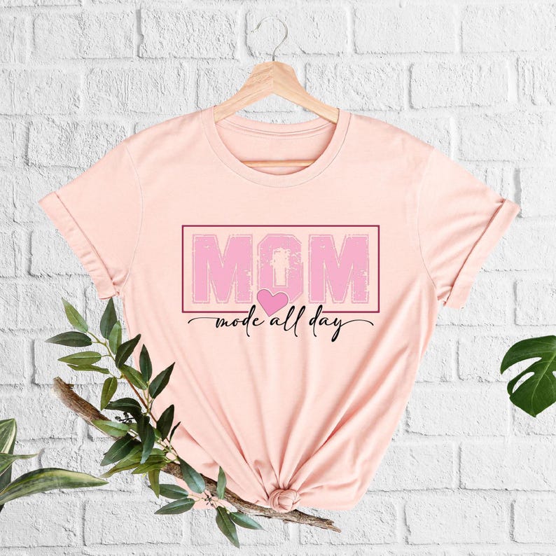 Puede incluir: Camiseta rosa claro con la palabra "MOM" en un recuadro rosa, un coraz&oacute;n rosa y la frase "mode all day" en cursiva. La camiseta est&aacute; colgada en una percha de madera sobre una pared de ladrillos blancos.