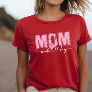 Puede incluir: Camiseta roja con la palabra "MOM" en rosa, con un dise&ntilde;o desgastado dentro de un rect&aacute;ngulo rosa. La frase "mode all day" est&aacute; escrita en cursiva debajo. La camiseta tiene mangas cortas.