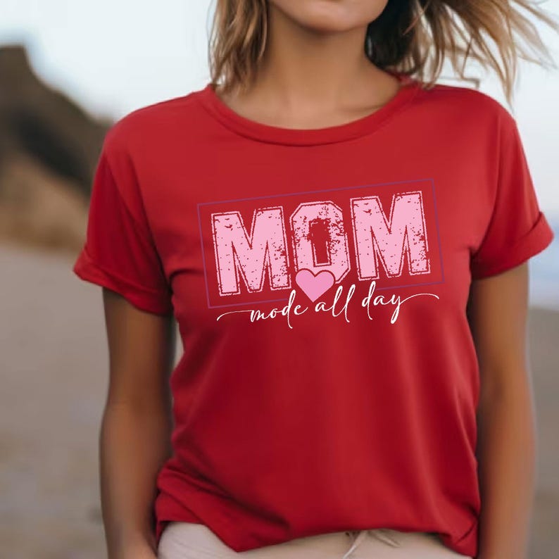Puede incluir: Camiseta roja con la palabra "MOM" en rosa, con un dise&ntilde;o desgastado dentro de un rect&aacute;ngulo rosa. La frase "mode all day" est&aacute; escrita en cursiva debajo. La camiseta tiene mangas cortas.