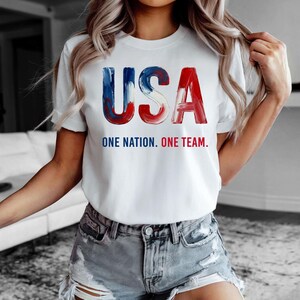 K&ouml;nnte beinhalten: Wei&szlig;es T-Shirt mit dem Wort "USA" in rot, wei&szlig; und blau. Unter dem Wort "USA" steht der Text "ONE NATION. ONE TEAM." Das T-Shirt hat einen Rundhalsausschnitt.