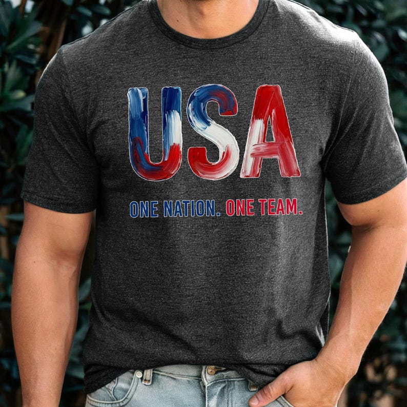 K&ouml;nnte beinhalten: Dunkelgraues T-Shirt mit dem Wort "USA" in rot, wei&szlig; und blau. Darunter steht der Text "ONE NATION. ONE TEAM."