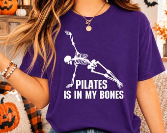 May include: A purple short-sleeve t-shirt featuring a white graphic of a skeleton performing a side plank exercise. Below the skeleton, the text reads "PILATES IS IN MY BONES" in bold white capital letters. The shirt has a crew neckline.
